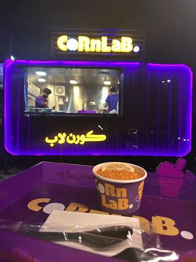 عربة CornLab ليلًا بإضاءة النيون، وكوب وصينية بالشعار في المقدمة